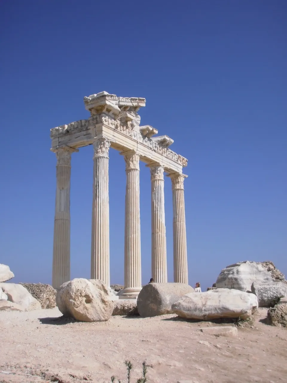 Looking up at the five Corinthian columns and their carved entablature against a deep blue sky, with fallen blocks in the foreground
