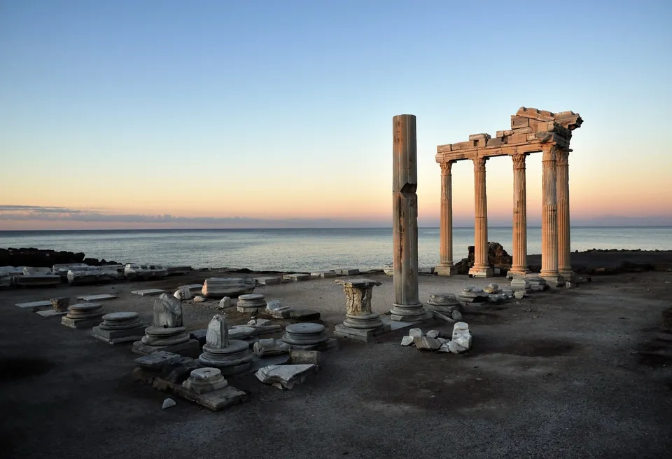 The Temple of Apollo at dusk: five lit columns against a pastel sky, scattered column bases and capitals on the dark platform, the sea flat behind